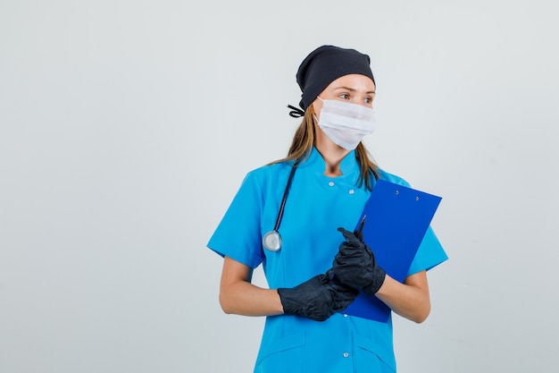 Kobieta lekarz w mundurze, rękawiczkach, masce patrząc na bok ze schowkiem i piórem