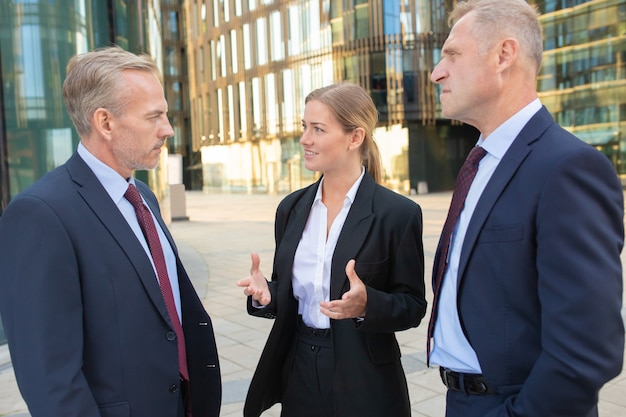 Bezpłatne zdjęcie kobieta menedżer omawiająca umowę z poważnymi klientami. biznesmeni w garniturach, stojąc i rozmawiając na zewnątrz z budynkiem miasta w tle. koncepcja spotkania i współpracy