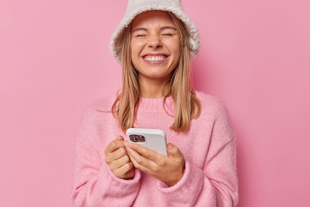 kobieta mruży oczy i uśmiecha się z radością, że otrzymała wiadomość, nosi swobodny sweter i czapkę zadowoloną z niesamowitej aplikacji na różowo