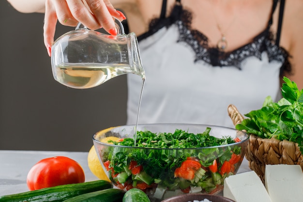 Kobieta nalewa oliwę z oliwek na sałatkę w szklanej misce z pomidorami, serem, zielenią, ogórkiem, widok z boku na szarej powierzchni