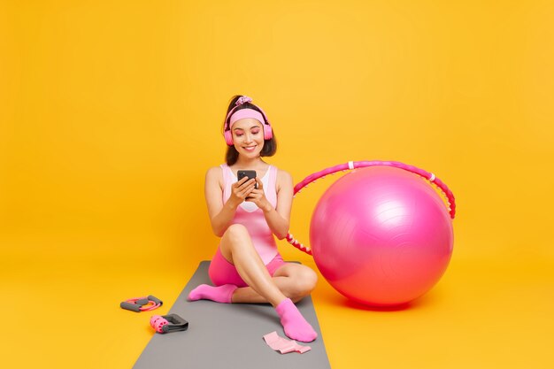 kobieta o ciemnych włosach sprawdza swoje osiągnięcia sportowe w aplikacji na smartfonie siada na macie fitness ubrana w odzież sportową używa piłki szwajcarskiej hula-hoop idzie do sportu pozuje w pomieszczeniu