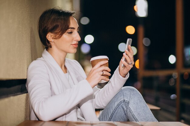 Kobieta opowiada na telefonie i pije kawę outside w ulicie przy nocą