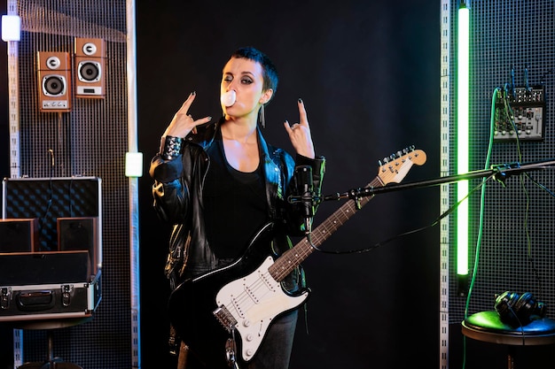 Kobieta performerka dmucha gumę balonową, pozując w studio, wykonując gest gwiazdy rocka, przygotowując gitarę elektryczną przed wykonaniem koncertu metalowego. Rebeliancki muzyk grający na występy solowe