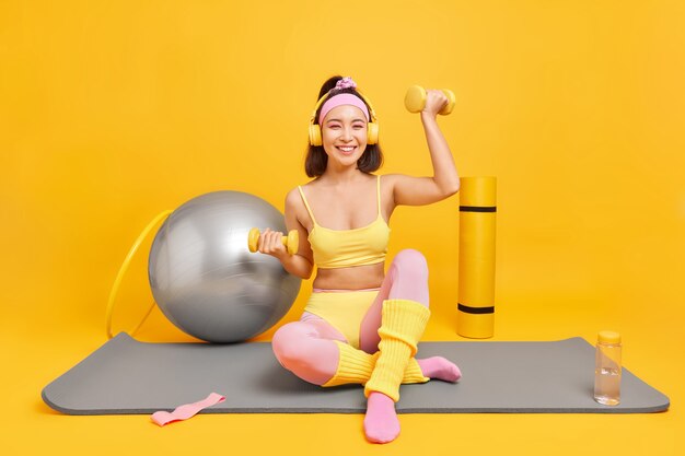 kobieta podnosi hantle słucha muzyki przez słuchawki nosi przycięty top legginsy z pałąkiem na głowę ma sportową figurę prowadzi aktywny tryb życia pozy na macie fitness na żółtym