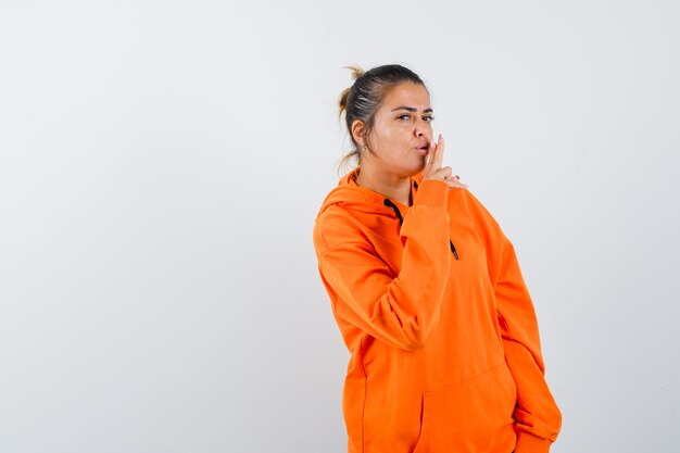 Kobieta pokazująca gest pistoletu w pomarańczowej bluzie z kapturem i wyglądająca na pewną siebie