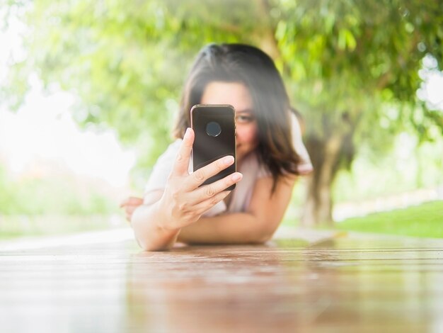 Kobieta położyć się na drewniany taras biorąc zdjęcie przy użyciu telefonu komórkowego w zielonym parku