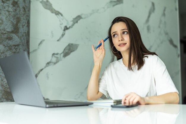 Kobieta pracuje na laptopie w domu w kuchni. pisanie w notatniku. Biznes kobieta myśli o czymś ważnym. Kobieta próbuje znaleźć pracę.