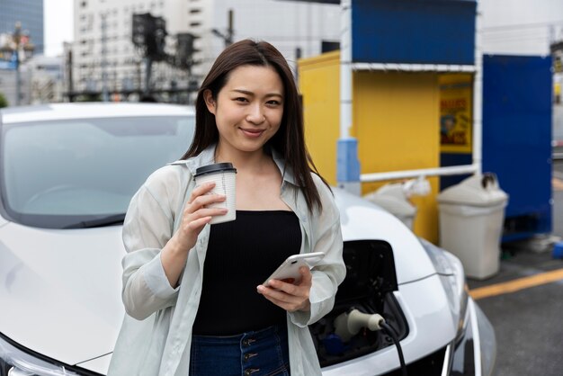 Kobieta robi sobie przerwę na kawę, gdy jej samochód elektryczny ładuje się i korzysta ze smartfona