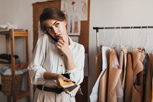 Kobieta rozmawiająca przez telefon, trzymająca próbki tkanin i w zamyśleniu pozująca