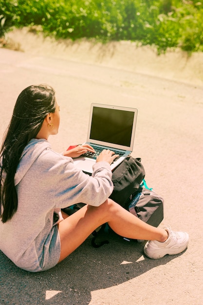 Bezpłatne zdjęcie kobieta siedzi na drodze i pracy na notebooku umieszczone na plecakach