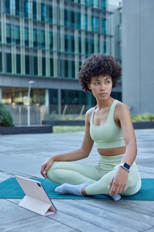 Kobieta siedzi ze skrzyżowanymi nogami na macie fitness ogląda samouczek na tablecie pozuje na zewnątrz ubrana w dres na tle nowoczesnego budynku robi ćwiczenia pilates