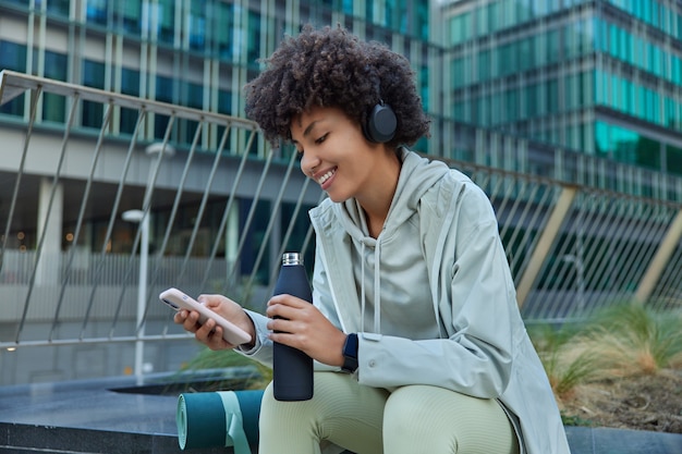 Kobieta słucha muzyki w słuchawkach przewija sieci społecznościowe za pomocą smartfona robi sobie przerwę po treningu pozuje na zewnątrz na tle nowoczesnego szklanego budynku będąc w dobrym nastroju