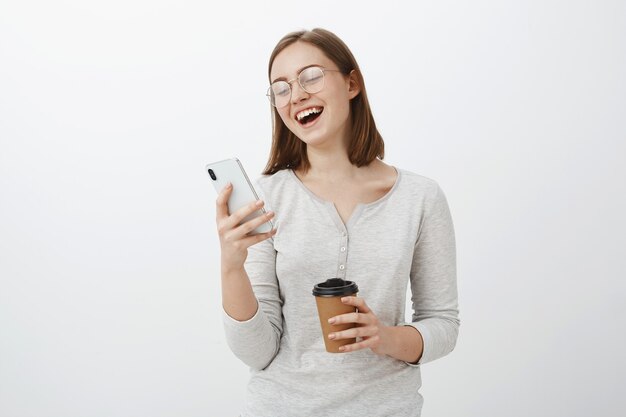 Kobieta śmiejąca się głośno, czytająca zabawny żart lub mem w Internecie, patrząc na ekran smartfona, trzymając papierowy kubek z kawą, bawiąc się, spędzając czas rozbawiony, czekając na przyjaciela w kawiarni