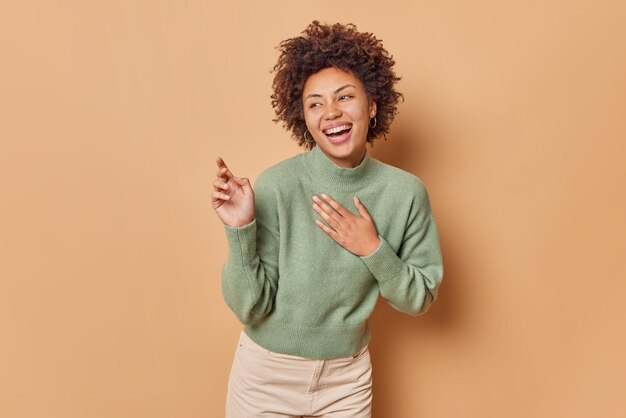 kobieta śmieje się i uśmiecha pokazuje szczere emocje trzyma rękę na klatce piersiowej odwraca wzrok chichocze na coś śmiesznego nosi swobodny sweter pozuje na tle beżowego studia. Szczęście