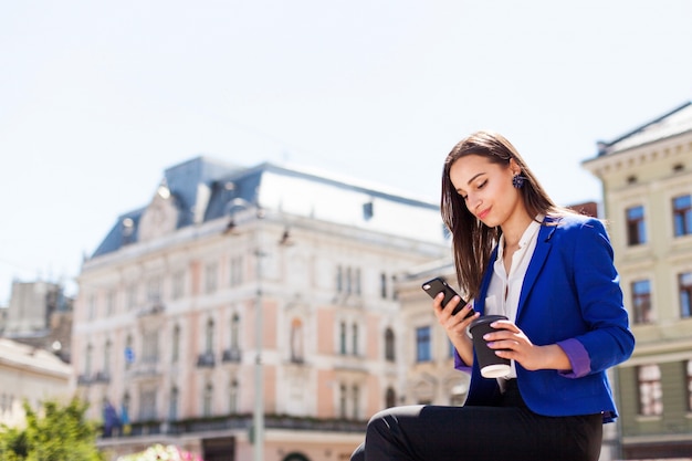 Kobieta Sprawdza Jej Telefonu Siedzi Z Filiżankę Kawy Na Ulicy