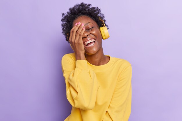 kobieta sprawia, że twarz uśmiecha się szeroko, śmieje się z czegoś bardzo zabawnego słucha muzyki przez słuchawki, ubrana w żółty sweter na fioletowo
