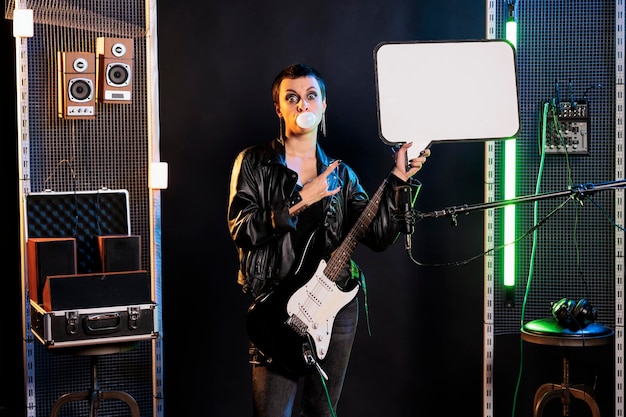 Bezpłatne zdjęcie kobieta supergwiazda wskazująca na pustą tablicę reklamową podczas gry na gitarze elektrycznej przygotowująca się do koncertu rockowego. zbuntowany muzyk w stylu grunge wykonujący heavy metalową piosenkę w studio