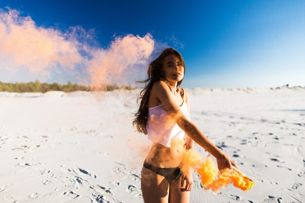 Kobieta tańczy pomarańczowy dym na białej plaży pod błękitnym niebem