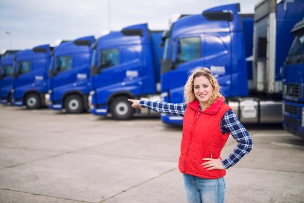 Kobieta trucker stojąca przed zaparkowanymi ciężarówkami i wskazująca palcem na pojazdy transportowe