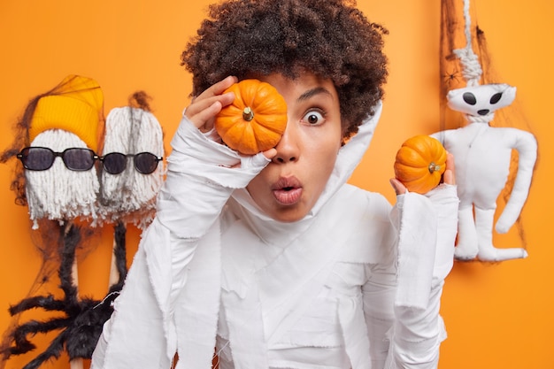 Bezpłatne zdjęcie kobieta trzyma dwie małe dynie patrzy zdziwiona nosi kostium białego ducha otoczonego halloweenowymi zabawkami na pomarańczowo