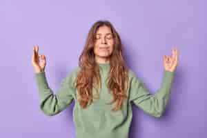 Bezpłatne zdjęcie kobieta trzyma ręce w geście zen om ma zamknięte oczy medytuje w pomieszczeniu swobodnie ubrana kontroluje emocje spokój i cierpliwość oddycha swobodnie na fioletowej ścianie