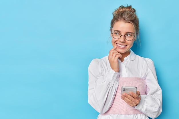 Kobieta Trzyma Telefon Komórkowy I Myśli O Czymś Przyjemnym Odwraca Wzrok, Nosi Okulary Optyczne I Białą Elegancką Koszulę Na Białym Tle Na Niebieskim Studiu. Użytkownik Technologii