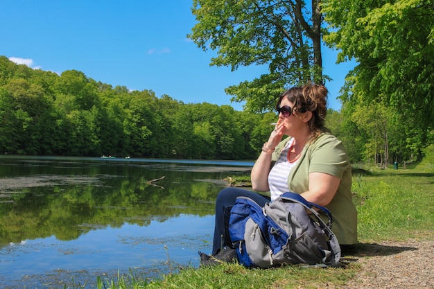 Bezpłatne zdjęcie kobieta turysta siedząca na brzegu jeziora z plecakiem i paląca papierosa