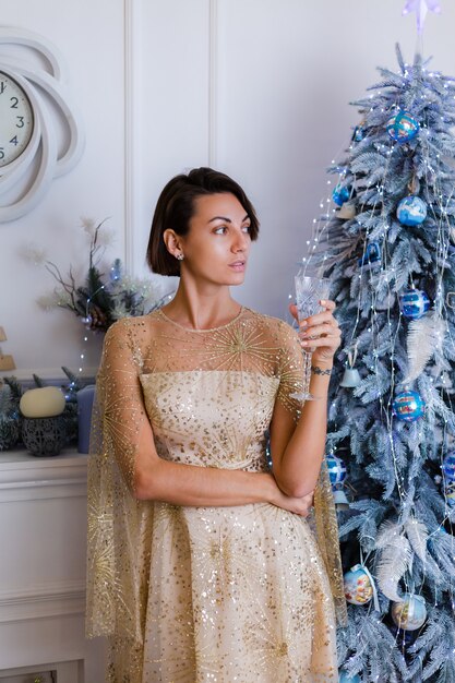 Kobieta ubrana w błyszczącą złotą suknię wieczorową boże narodzenie trzyma kieliszek szampana przez niebiesko-zielone drzewo roku w domu