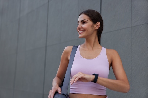 kobieta ubrana w strój sportowy ma na sobie smartwatch niosący zwinięty karemat odwraca wzrok z delikatnymi uśmiechami przy szarej ścianie będąc w dobrym nastroju