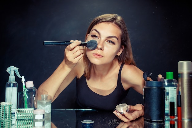 Bezpłatne zdjęcie kobieta uroda stosowania makijażu. piękna dziewczyna patrzeje w lustrze i stosuje kosmetyka z dużym muśnięciem.