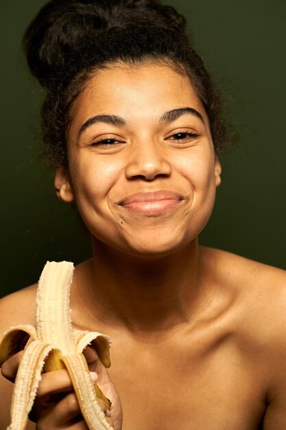 Bezpłatne zdjęcie kobieta uśmiechająca się do kamery, trzymająca i jedząca dojrzałego żółtego banana odizolowanego na zielono