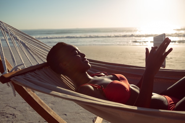 Kobieta używa cyfrową pastylkę podczas gdy relaksujący w hamaku na plaży