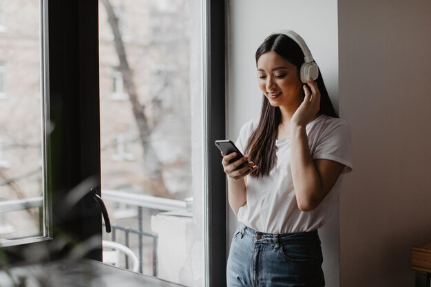 Kobieta w białej koszulce i dżinsach pozuje przy oknie z telefonem w dłoniach Dziewczyna słucha muzyki na słuchawkach