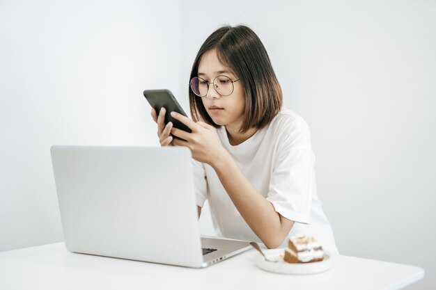 Kobieta w białej koszuli, grająca w smartfona i posiadająca laptopa.