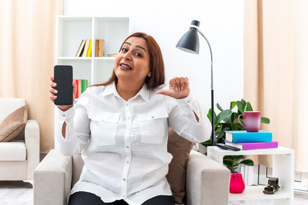 Kobieta w białej koszuli i czarnych spodniach pokazująca smartfona wskazującego palcem wskazującym uśmiechnięta siedząca na krześle w jasnym salonie