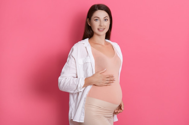 Kobieta w ciąży pieści brzuch patrząc na kamery, ciemnowłosa przyszła mama pozuje na różowej ścianie, dotyka jej brzucha, ma pozytywne emocje.