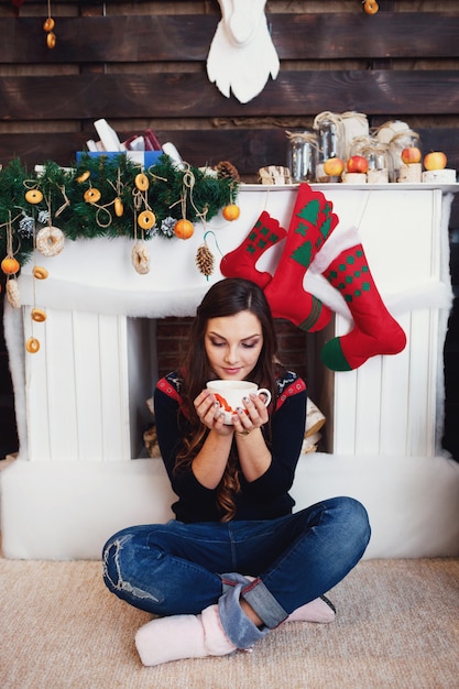 Kobieta w dżinsach siedzi z filiżanką gorącego napoju przed kominkiem ozdobionym świątecznymi rzeczami