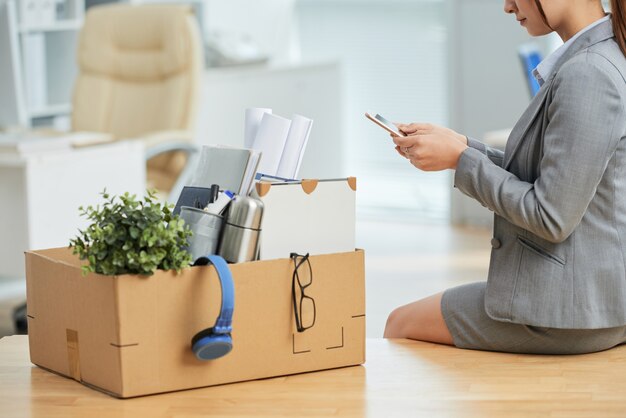 Kobieta w garniturze, siedząc na biurku w biurze z rzeczy w pudełku i przy użyciu smartfona