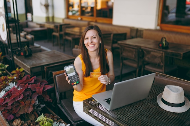 Kobieta w kawiarni na świeżym powietrzu siedząc z laptopem na komputerze, trzyma bezprzewodowy terminal płatniczy nowoczesny bank do przetwarzania płatności kartą kredytową