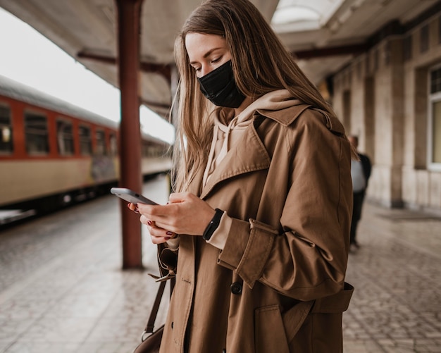 Kobieta w masce i przy użyciu telefonu komórkowego na stacji kolejowej