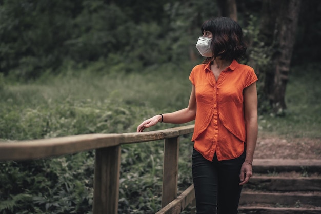 Kobieta w masce na twarz stojąca na drewnianym moście w lesie w dzień - COVID-19