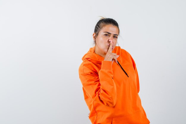 Kobieta w pomarańczowej bluzie z kapturem pokazująca gest ciszy i wyglądająca rozsądnie