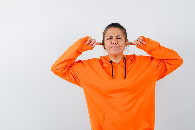 Kobieta w pomarańczowej bluzie z kapturem zatykająca uszy palcami i wyglądająca na zirytowaną