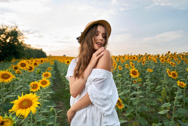 Kobieta w słomkowym kapeluszu i białej sukni pozuje na słonecznikowym polu
