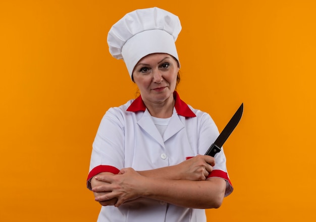 kobieta w średnim wieku kucharz w mundurze szefa kuchni skrzyżowaniu rąk trzymając nóż na izolowanych żółtej ścianie z miejsca na kopię
