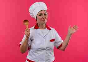 Bezpłatne zdjęcie kobieta w średnim wieku kucharz w mundurze szefa kuchni trzymając łyżkę rozprzestrzeniać rękę na izolowanej ścianie różowy