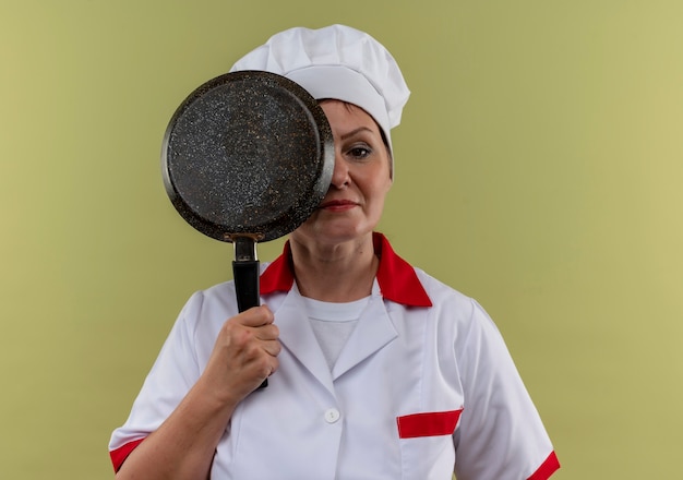 Bezpłatne zdjęcie kobieta w średnim wieku kucharz w mundurze szefa kuchni zakryte oko z patelni na odizolowanej zielonej ścianie