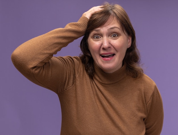 Kobieta w średnim wieku w brązowym golfie mylić z ręką na głowie za pomyłkę stojąc nad fioletową ścianą