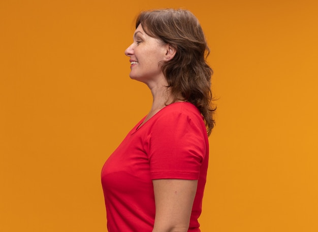 Kobieta w średnim wieku w czerwonej koszulce stojącej bokiem z uśmiechem na twarzy na pomarańczowej ścianie