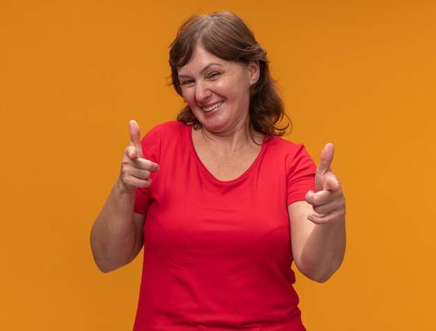 Kobieta w średnim wieku w czerwonej koszulce, wskazując palcami wskazującymi, szczęśliwa i wesoła stojąca nad pomarańczową ścianą